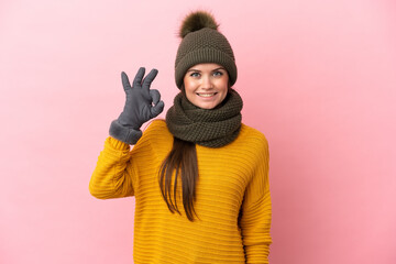 Young caucasian girl with winter hat isolated on pink background showing ok sign with fingers