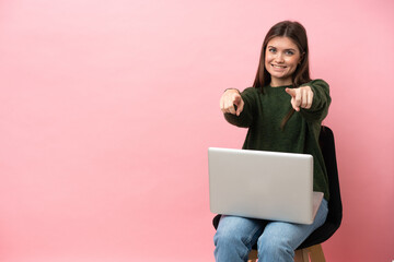 Young caucasian woman sitting on a chair with her laptop isolated on pink background points finger at you while smiling - 790669205