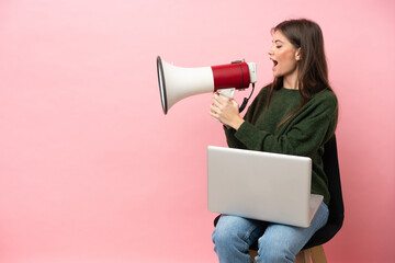 Young caucasian woman sitting on a chair with her laptop isolated on pink background shouting through a megaphone - 790669215