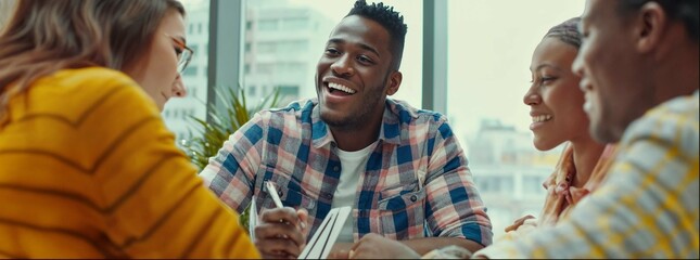 A group of young people in an office, smiling and having fun while they study together with books on the table. A black man wearing a checkered shirt is talking to his friends, all dressed casually. 