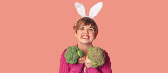 Studio shot of a young Caucasian woman in bunny ears holding fresh broccoli in her hands. healthy...