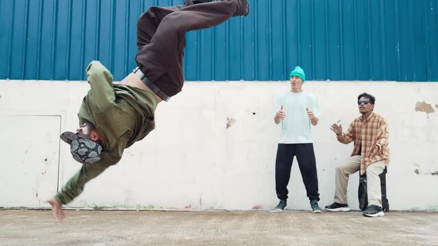 Skilled hipster doing cart wheel while multicultural friends encourage him. Hip-hop dancer moving foot step to music surrounded by group of hipster at wall near street. Outdoor sport 2024. hiphop.