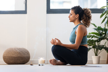 Sporty young woman doing yoga and hypopressive exercises while staying in lotus position in living room at home. - obrazy, fototapety, plakaty