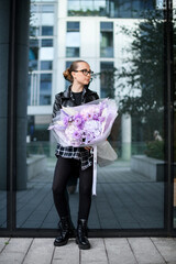 Cute slender woman holds a large bouquet of purple flowers against the background of the glass mirror wall of the building