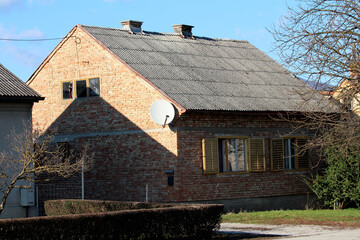 Red brick small old unfinished urban family house with yellow dilapidated wooden window blinds and without facade next to tall trees without leaves