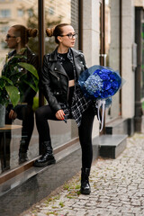Young pretty woman standing near a glass shop window holding a bouquet of blue hydrangeas and blue...