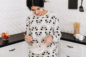 Cheerful woman standing in bright kitchen mixing dough in a bowl, making homemade cookies, dressed...