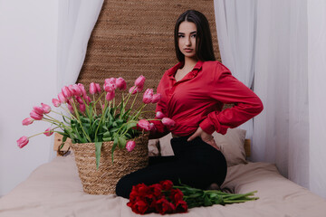 A woman poses lying on bed, surrounded by vibrant bouquets bunch of red tulips placed around the room, suggesting a serene and domestic atmosphere. Spring International Women's Day, 8th of March
