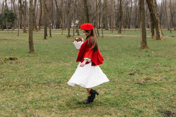 Young woman 20s in red french beret and jacket dressed white dress, walking in city park with...