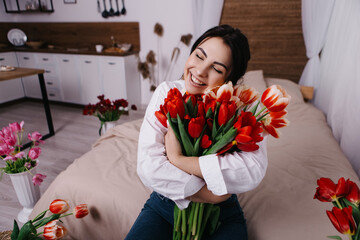 A woman poses on bed, surrounded by vibrant fresh bouquets bunch of red tulips placed around the...