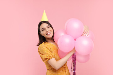 Happy young woman in party hat with balloons on pink background