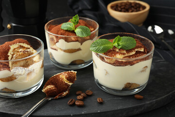 Delicious tiramisu in glasses, spoon, coffee beans and mint leaves on table