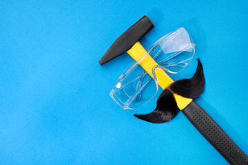 Man's face made of artificial mustache, safety glasses and hammer on blue background, top view....