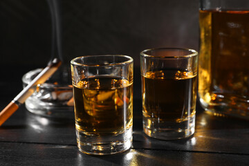 Alcohol addiction. Whiskey in glasses, smoldering cigarette and ashtray on black wooden table,...