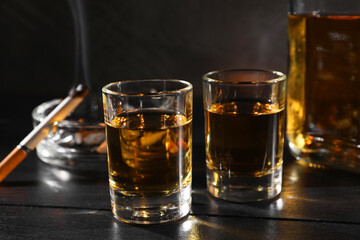Alcohol addiction. Whiskey in glasses, smoldering cigarette and ashtray on black wooden table, closeup