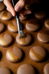 Female confectioner's hand paints brown macaroons with golden decorative pastry paint