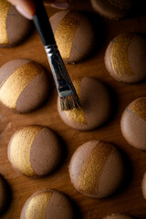 Female hand draws golden stripes on brown macaroons with paint for confectionery products