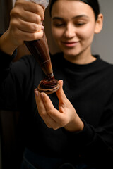 Smiling young woman holds a half of a brown macaroon in front of her, on which she applies chocolate cream