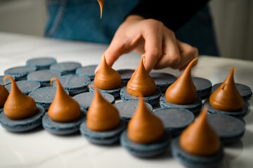 Close-up photo of blue macaroon halves on which a female confectioner squeezes chocolate cream