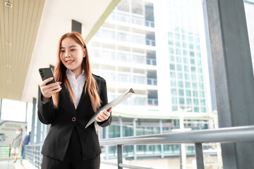 Fototapeta premium Happy young Asian female businesswoman using cell phone smartphone and holding document files phone at the walkway