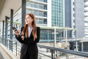 Fototapeta premium Happy young Asian female businesswoman using cell phone smartphone and holding document files phone at the walkway