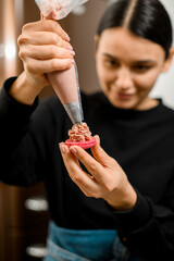 Focus on the red half of the macaroon, on which the female pastry chef applies pink cream from a pastry bag