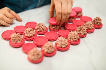 Red pieces of macaroons lie on a white kitchen table, one of which has beige cream in a curly shape