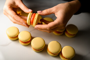 Female hands of a confectioner fasten a beige macaroon smeared with pink cream, holding it directly above the table