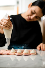 Female hands of a confectioner squeeze white cream from a pastry bag onto pink halves of macaroons
