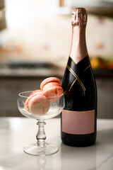Tall glass with pink macaroons stands on the kitchen table, a bottle of champagne stands next to it