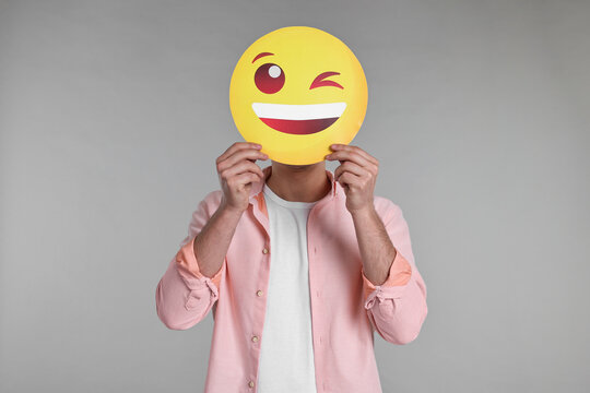 Man covering face with happy winking emoticon on grey background - Powered by Adobe