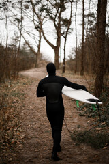 Young male surfer wearing a wetsuit walks along a path between the trees, holding his surfboard