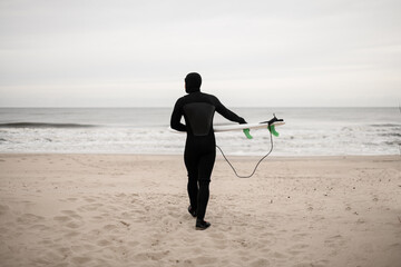 Young male surfer wearing a wetsuit is walking on the sand, holding his surfboard