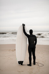 Male surfer in black wetsuit standing on sandy seashore with white surfboard standing on sand