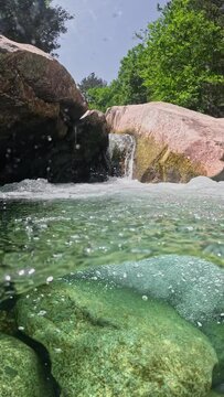 Half underwater slow motion shot of fast mountain stream with crystal clear water. River water flows between the stones forming small waterfalls and baths, VERTICAL VIDEO 