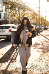 Young pretty smiling woman walking on the sidewalk holding a disposable cup with coffee