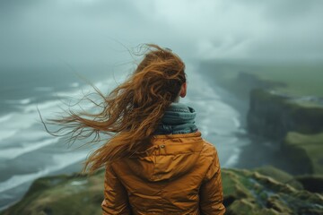 Woman in orange jacket and scarf stands with windblown hair facing a stormy sea and rugged coastline