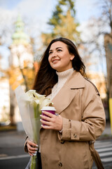 Young pretty smiling woman with long hair, holding a bouquet of lilies and a disposable cup of coffee
