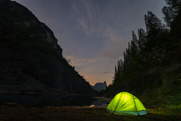 A yellow tent sits next to a large pond in the mine shaft. There is a pine forest next to the pond and a large mountain on the other side..Star shooting adventure In the dark night.water reflection