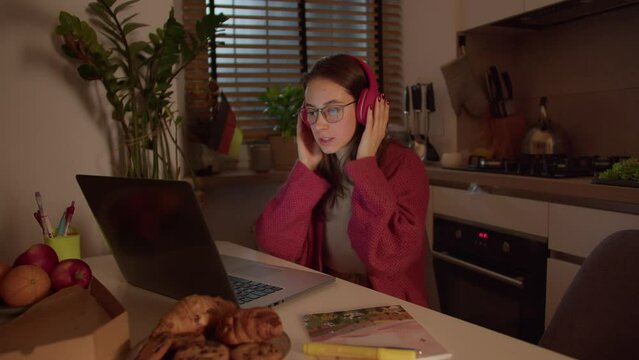A confident brunette girl in red wireless headphones and glasses listens to the correct pronunciation of the native speaker and studies foreign languages online at home in the kitchen in front of a