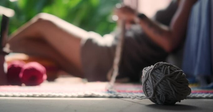 In the foreground is a ball of woolen thread in the background out of focus is a young woman with a short haircut who sits on the floor and knits a blanket with a basket of tools standing next to her.