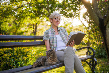 Happy senior woman enjoys reading book and spending time with her cat while sitting on a bench in her garden. - 790819619