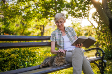 Happy senior woman enjoys reading book and spending time with her cat while sitting on a bench in her garden.