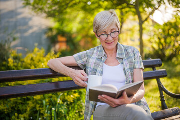 Happy senior woman enjoys reading book and drinking coffee on a bench in her garden.