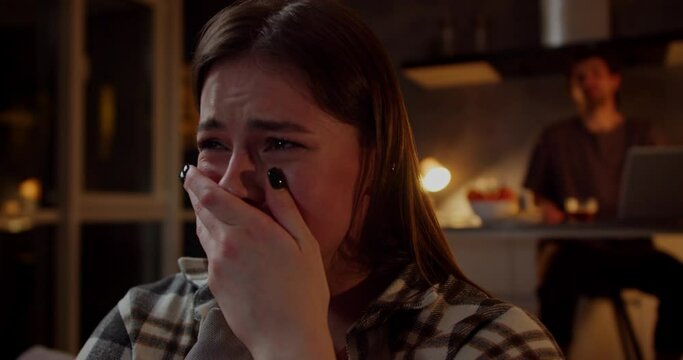 A brunette girl in a plaid shirt cries while watching a sad drama on TV and her brunette boyfriend in a gray T-shirt consoles the girl and hugs her in a modern studio apartment in the evening