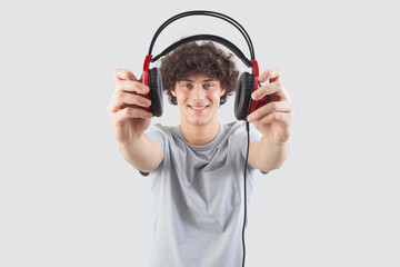 Young handsome smiling man, holding headphones in his hand and showing them to the camera before putting them on to listen to music or play video games, isolated against a gray background