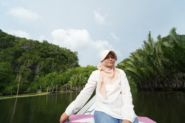 Fototapeta premium Portrait of woman sitting in wooden boat. Asian woman wearing a hijab sits on a wooden boat that is traveling along a river with mangrove trees around it