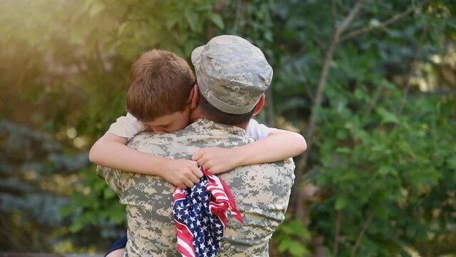 A soldier father is hugging his son holding an American flag outside for a family reunion or 4th of July concept.