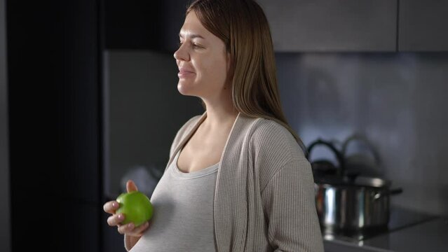 A happy pregnant woman takes a green apple and deftly tosses it in her hand, sniffs it, enjoying its aroma. Thoughtful woman holding an apple in her hand and gently touching her belly