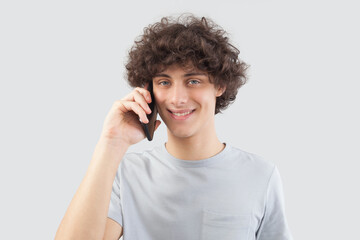 A young, handsome man smiles as he uses his smartphone, talking and listening with cellphone while looking into the camera with his blue eyes. He is isolated against a gray background